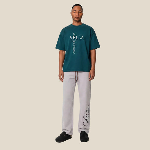 Man wearing a teal Vella t-shirt and gray pants on a white background
